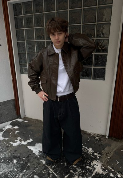 Chaqueta bomber de cuero marrón con pantalones amplios de mezclilla. Polo blanco debajo, grandes anillos de plata. De pie en un entorno al aire libre nevado.