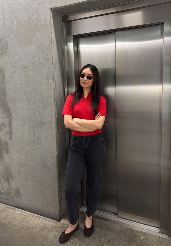 Jeune femme en chemise rouge et jean noir se tient bras croisés près des portes fermées d’un ascenseur en acier inoxydable.