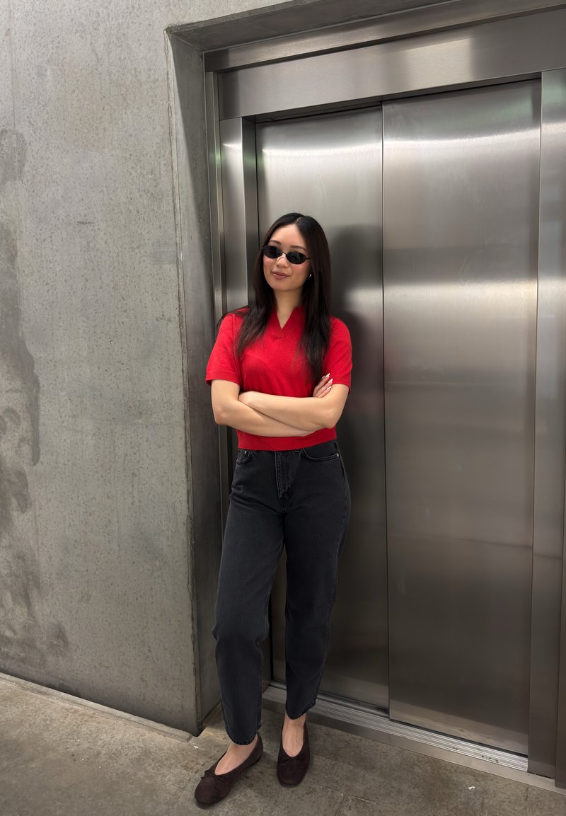 Young woman in red shirt and black jeans stands with arms crossed by closed stainless steel elevator doors.