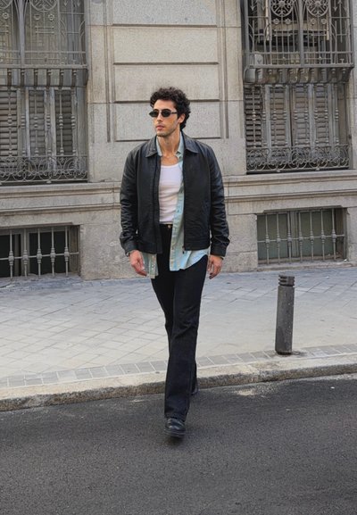 Hombre con chaqueta de cuero negra, camiseta blanca sin mangas, camisa azul claro, pantalones negros y gafas de sol caminando por una calle de la ciudad.