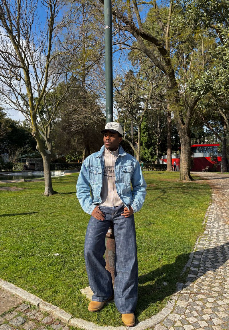 Jeune homme portant une veste en jean claire, un jean large, un pull beige, une casquette et des bottes, appuyé contre un lampadaire dans un parc ensoleillé avec des arbres et un chemin pavé.