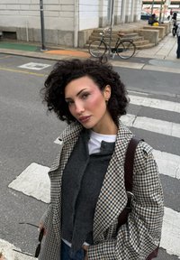 Rutigt ullkappa i beige och marinblått med vida ärmar, lager-på-lager över en grå stickad tröja. Tillbehör inkluderar en brun väska och svarta solglasögon.