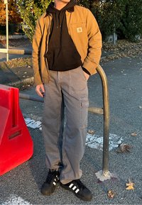 Brun arbejdsjakke over en sort hoodie, grå cargo-bukser og sorte sneakers med hvide striber. Står ved siden af en rød barriere på et brolagt område.