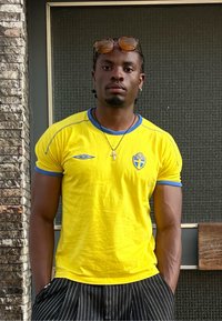 Κίτρινη αθλητική φανέλα με μπλε λεπτομέρειες, διαθέτει κυκλικό λαιμό, κεντητό λογότυπο και κοντά μανίκια. Μαύρο κοντό παντελόνι με ρίγες.