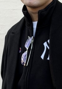 Černá bunda na zip s výrazným vyšitým logem, nošená přes bílou košili. Stříbrné sluneční brýle s reflexními čočkami visí na zipu.