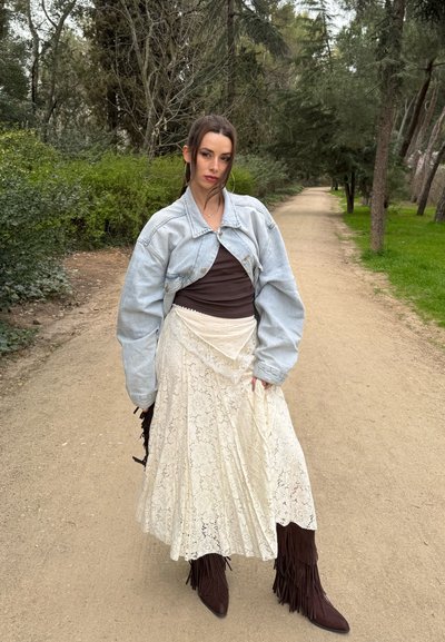 Mujer de pie en un sendero del bosque con una chaqueta de mezclilla clara, camiseta oscura, falda blanca de encaje y botas marrones con flecos, rodeada de árboles y vegetación.