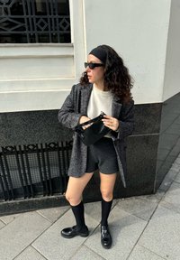 Abrigo de tweed gris sobre un suéter blanco, pantalones cortos negros y calcetas negras hasta la rodilla. Los accesorios incluyen un bolso negro y gafas de sol.