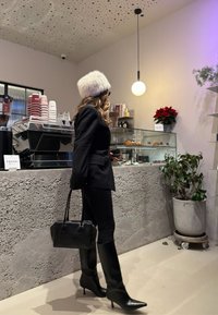 Blazer noir sur mesure, pantalon noir ajusté, bottes à talons aiguilles noires montant au genou, et un chapeau en fausse fourrure blanc. Sac à main noir, intérieur de café.