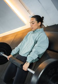 Ljusgrönblå vindjacka med elastiska muddar och vita detaljer, buren av en person som lyfter en skivstång med svart vadderat grepp i en gymmiljö.