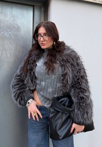 Veste en fausse fourrure grise avec un col montant, pull gris côtelé avec des motifs d'étoiles, jean bleu et sac en cuir noir à la main.