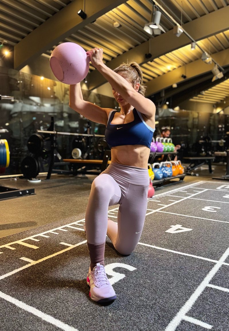 Kvinna i träningskläder knäböjer på ett knä i ett gym och lyfter en rosa kettlebell ovanför huvudet, med gymutrustning synlig i bakgrunden.
