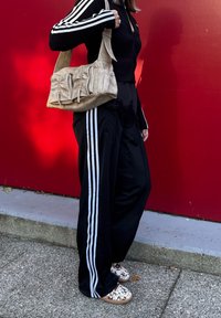 Črne hlače z belimi stranskimi črtami, črn zip-up zgoraj in bež torba v stilu slouchy. Rjav in bel vzorčasti čevlji na zavezovanje. Rdeča ozadje.