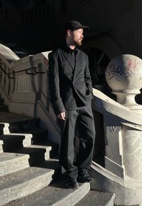 Costume à fines rayures noires avec une veste ajustée et un pantalon ample. Porté avec une chemise noire et une casquette ; debout sur un escalier avec des graffitis.