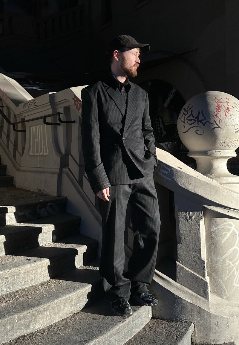 Costume à fines rayures noires avec une veste ajustée et un pantalon ample. Porté avec une chemise noire et une casquette ; debout sur un escalier avec des graffitis.