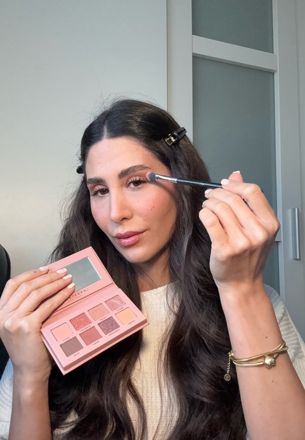 Una palette rettangolare di ombretti rosa con nove tonalità, inclusi finish opachi e glitterati, tenuta in una mano. Nell'altra mano un pennello per trucco.