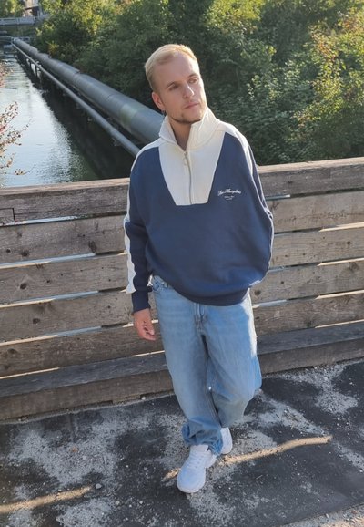 Sudadera con cremallera en azul marino y blanco, con un alto cuello, combinada con jeans holgados de color azul claro y zapatillas blancas, de pie sobre una plataforma de madera.