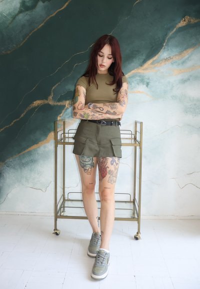 Top ajustado de color verde oliva combinado con una falda a juego que cuenta con dos bolsillos frontales. Zapatillas grises y tatuajes intrincados en las piernas visibles. Fondo con toques dorados.