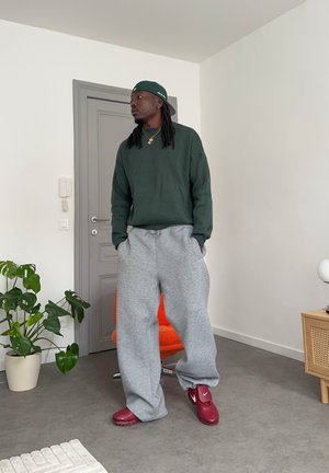 Hombre con sudadera verde, pantalones deportivos grises, zapatillas rojas y gorra al revés, de pie con las manos en los bolsillos, en interiores, cerca de una planta y una silla naranja.