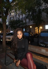 Μαύρη μπλούζα με μακριά μανίκια, παγιέτα φούστα και μπορντό κολάν. Κάθεται σε μια ξύλινη παγκάκι με θολό αστικό φόντο και μαλακό φωτισμό.