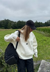 Felpa bianca con un logo nero triangolare, abbinata a pantaloni neri e un cappellino nero. Una borsa nera con una tasca in rete è indossata a spalla.