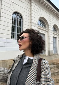 Rutig kappa med mörkgrå tröja, svarta solglasögon med guldaccenter. Lockigt hår, profilvy mot en vit byggnad.