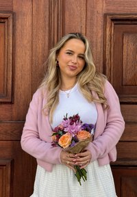 Blonde Frau in einem pinkfarbenen Cardigan, die einen bunten Strauß aus orangefarbenen Rosen, lila Blumen und Grünpflanzen hält, vor einer Holztür.