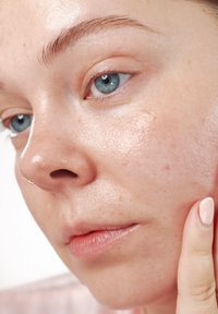 Zbliżenie na twarz o jasnej karnacji, niebieskich oczach i lekkich piegach. Skóra wygląda na gładką i błyszczącą, a palec delikatnie dotyka policzka.