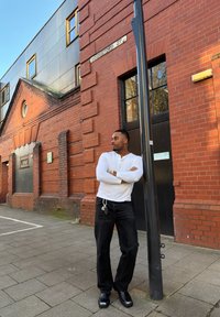 Homme en chemise blanche à manches longues et pantalon noir appuyé contre un poteau noir sur un trottoir en briques au coin d'une rue.