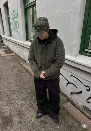 Persona con gorra de camuflaje y chaqueta verde, de pie en una calle junto a una pared blanca con ventanas con marco verde y graffiti.