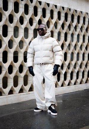Chaqueta blanca almohadillada, pantalones blancos, guantes negros y zapatos negros con suelas blancas gruesas, contra un fondo de pared geométrica texturizada.