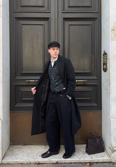 Hombre con abrigo negro de inspiración vintage, chaleco azul marino, pantalones de pierna ancha y gorra, de pie frente a una gran puerta de madera oscura junto a un bolso de cuero tejido.