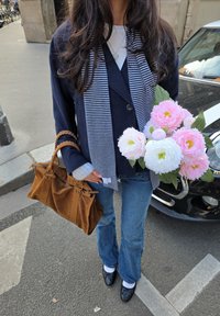 Barna velúr kézitáska, tengerészkék blézer csíkos sállal, kék farmernadrág, fehér zokni, fekete cipő, rózsaszín és fehér virágokból álló csokrot tartva.