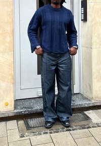 Personne portant un pull bleu texturé, un jean à jambes larges et des chaussures noires, debout sur un trottoir carrelé devant une porte blanche.