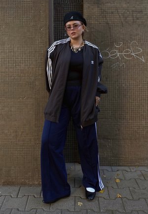 Chaqueta atlética negra con rayas blancas, parte superior negra, pantalones de pierna ancha azul marino, boina negra, gafas de sol rosadas, collar de declaración dorado.