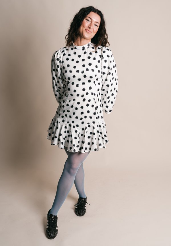 Femme debout, jambes croisées, portant une robe blanche à pois noirs, des collants bleus et des baskets noires, souriant doucement.