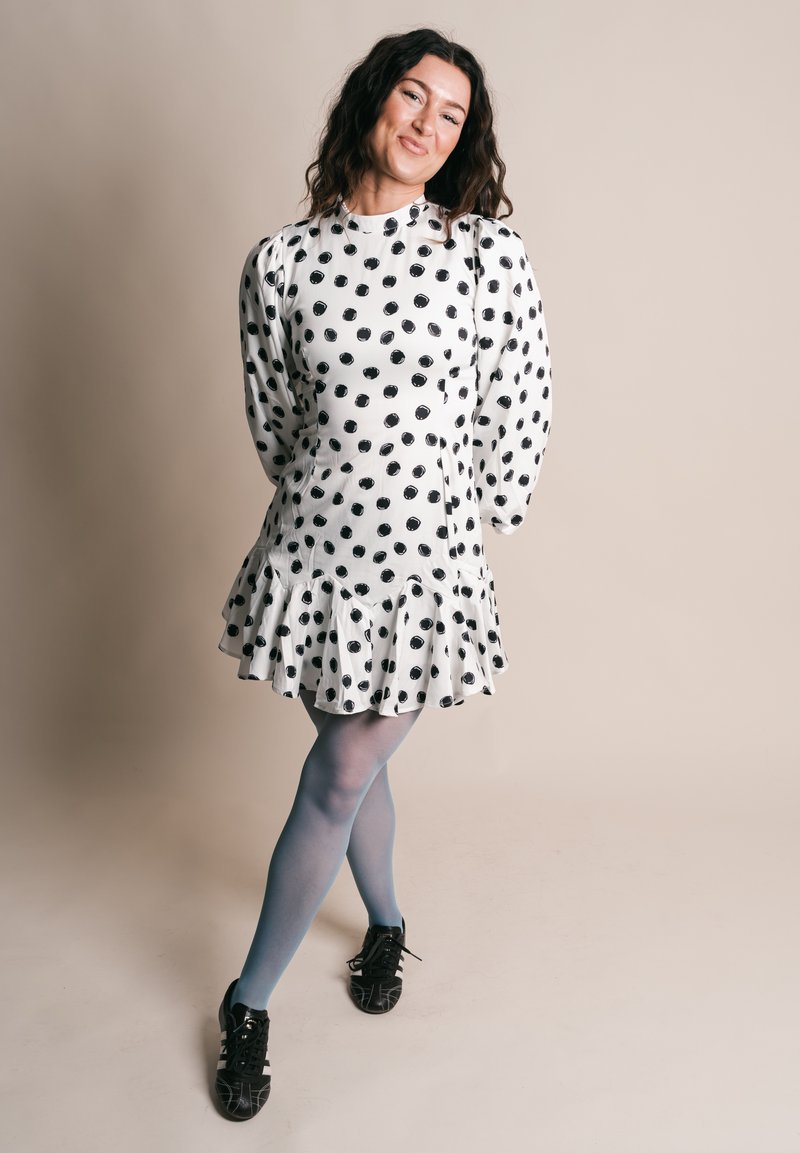 Egy nő áll, keresztbe tett lábakkal, fehér ruhát visel fekete pöttyökkel, kék harisnyát és fekete tornacipőt, finoman mosolyogva.