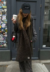 Galvā pelēkas krāsas topu un brūnu leoparda printa šalli, rokās turot kafiju. Melna cepure un raiba bikses. Melna plecu soma ar sarkanām akcentiem.