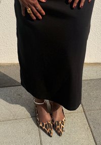 Leopard print high-heeled shoes with pointed toes, teamed with a black skirt, set against a stone pavement. A delicate beaded bracelet adorns the ankle.