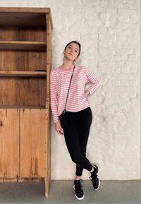 Roosa ja valge triibuline pikkade varrukatega särk, mustad teksad, mustad tossud ja väike üle õla kott, poseerides puidust riiuli taustal.