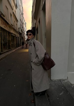 Abrigo largo gris con botones negros, lleva una boina negra y gafas de sol, sosteniendo un bolso marrón de hombro en una calle estrecha al anochecer.