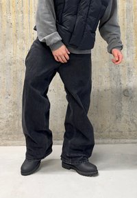 Jeans neri a gamba ampia con una texture sbiadita, abbinati a una felpa grigia e a un gilet imbottito nero. Le calzature sono nere, in gomma e robuste.