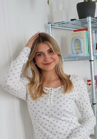 Camisa blanca de manga larga con estampado de lazos azul claro, presenta un escote redondo con botones. Tela suave y ajuste cómodo visibles.