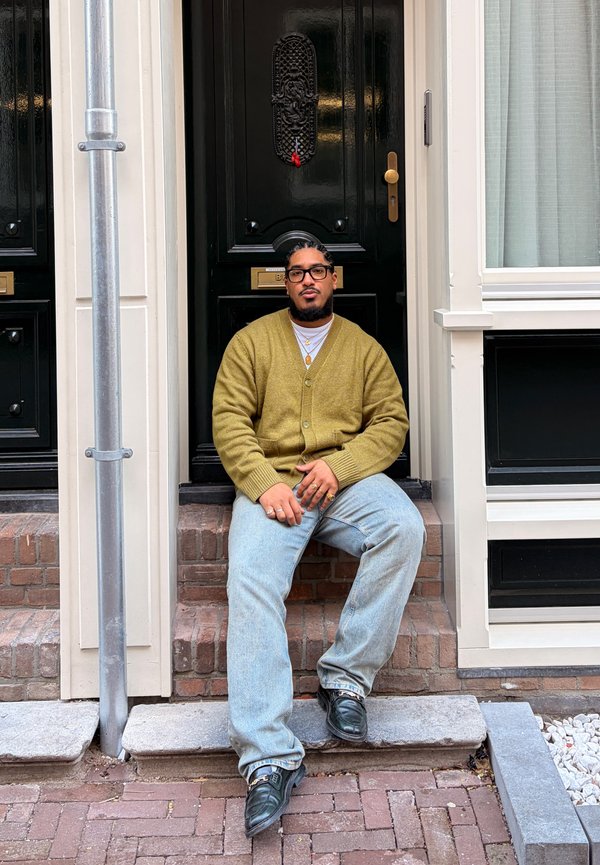 Mann mit Brille, grünem Cardigan und hellen Jeans, sitzt auf Ziegelstufen vor einer dunklen Tür mit einem Messing-Briefschlitz.