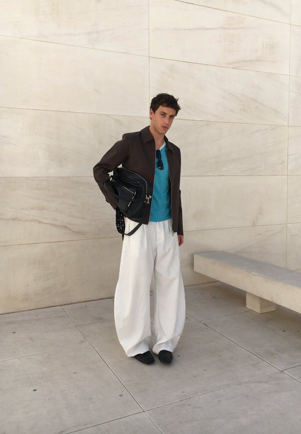 Jeune homme portant une veste noire, une chemise bleue, un pantalon large blanc, tenant un sac noir, debout contre un mur de pierre clair près d'un banc.