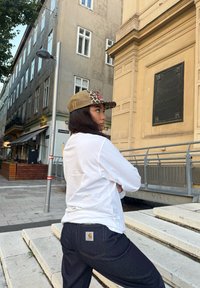 Valge pika varrukaga särk, tumedad teksad Carhartt sildiga ja leopardimustriga müts, millel on võrkmaterjal tagant, linnatausta ees.