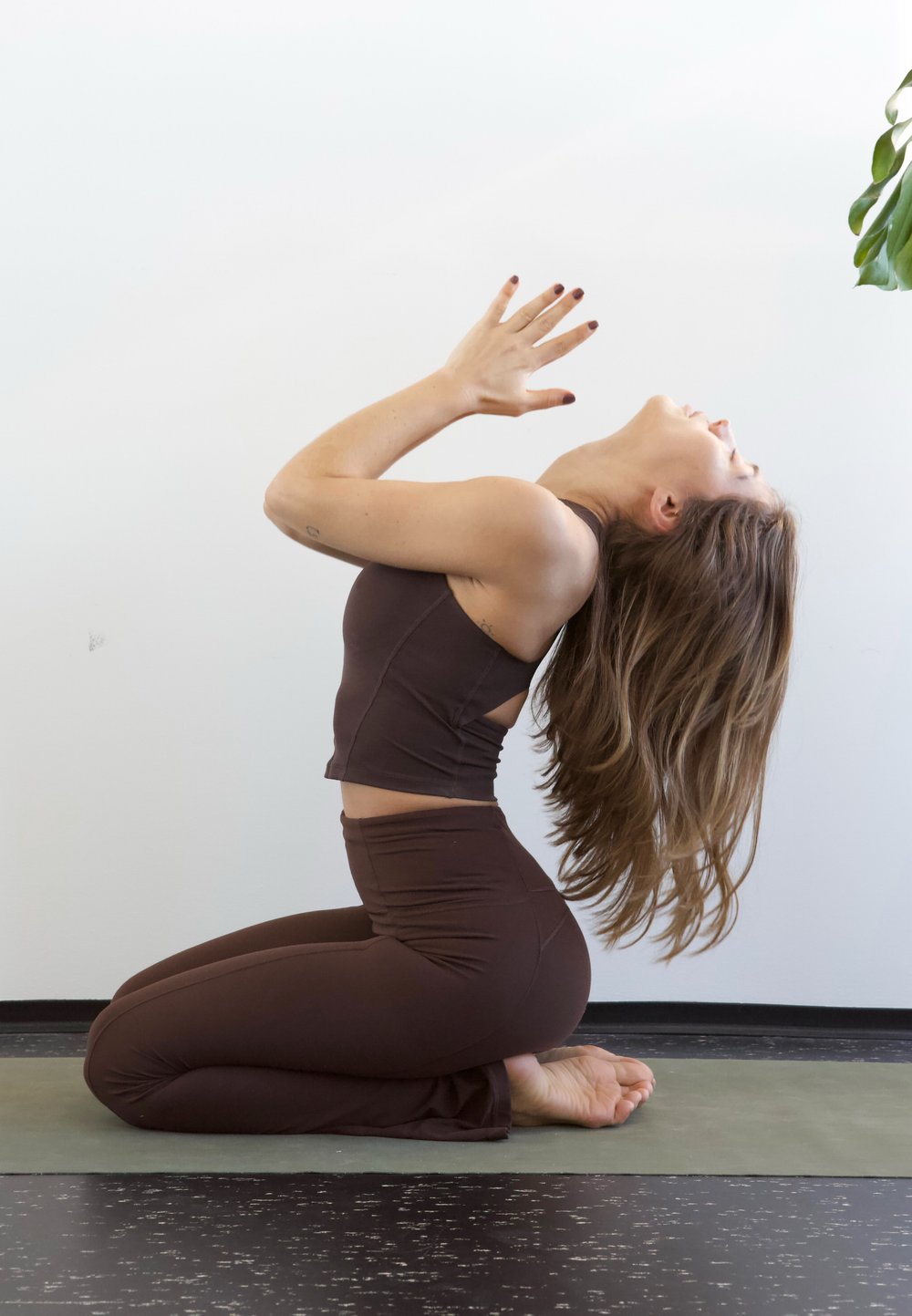Top de desporto castanho e leggings, de joelhos numa esteira de ioga verde. Cabelo comprido a fluir, mãos levantadas numa posição de oração contra um fundo claro.