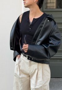 Persona che indossa una giacca di pelle nera sulle spalle, una maglietta senza maniche nera, pantaloni plissettati crema, tenendo degli occhiali da sole in una mano.