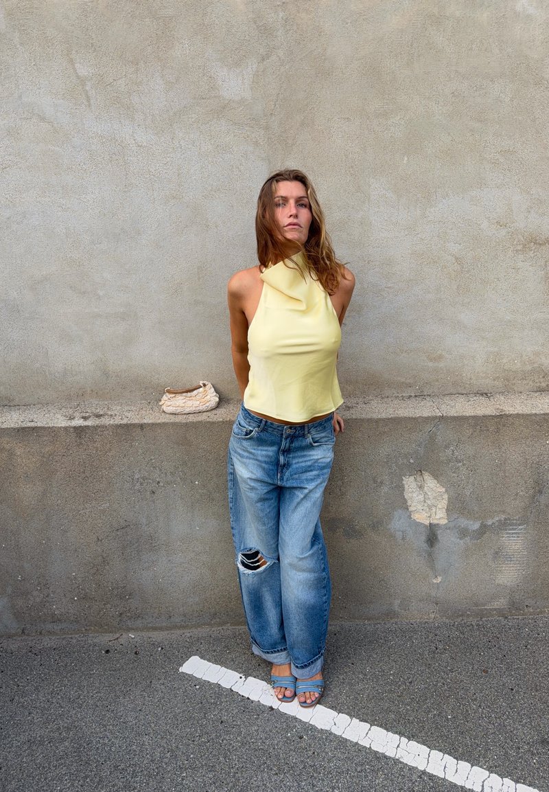 Femme en top jaune à col halter et jean bleu déchiré se tient contre un mur gris texturé avec un petit sac blanc à côté d'elle.
