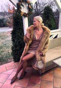 Lang, fuskepelsjakke i beige med krage av myk pels over en beige kjole, kombinert med spisse hæler og en liten sølvfarget veske. Utendørs setting.