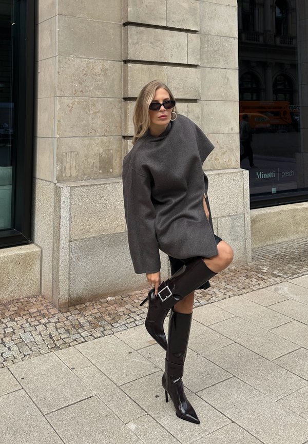 Maglione grigio oversize in lana con collo alto, abbinato a stivali neri in pelle lucida con tacco a stiletto che presentano un dettaglio con fibbia.
